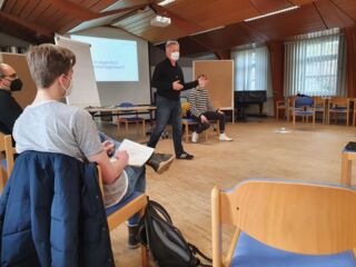 Guido Froese steht in der Mitte von einem Stuhlkreis mit den Teilnehmer*innen des MUSIK21-Kurses und erklärt etwas.