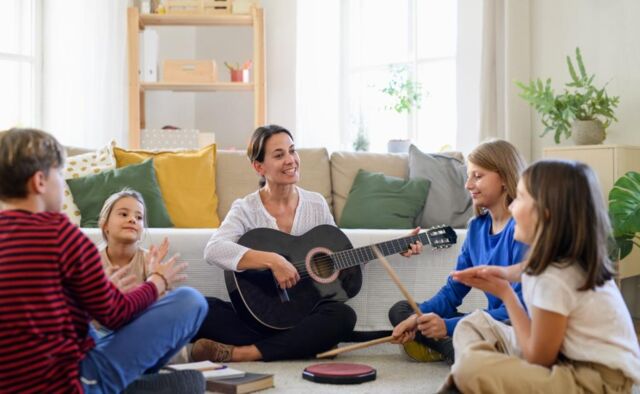 Frau sitzt mit einer Gruppe von Kindern auf dem Boden und spielt Gitarre, während die Kinder dazu einen Rythmus schlagen