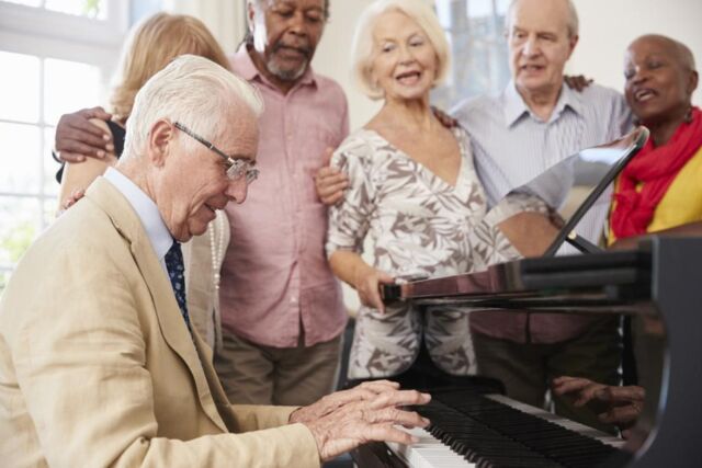 5 ältere Menschen stehen Arm in Arm um einen Flügel und singen. Einer sitzt am Klavier und begleitet.