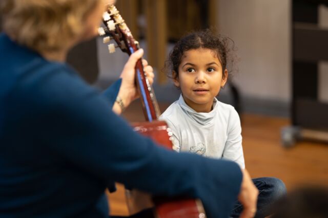 Ein Kind hört zu, wie ein Mensch (nur verschwommen von hinten zu sehen) Gitarre spielt.
