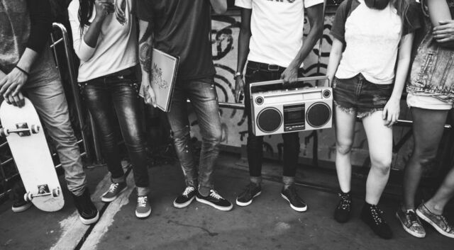 Bildausschnitt von 6 Jugendlichen, welche in lässiger Haltung nebeneinander stehen und unter anderem ein Skateboard oder ein Radio in der Hand halten. Das Foto ist schwarz-weiß.